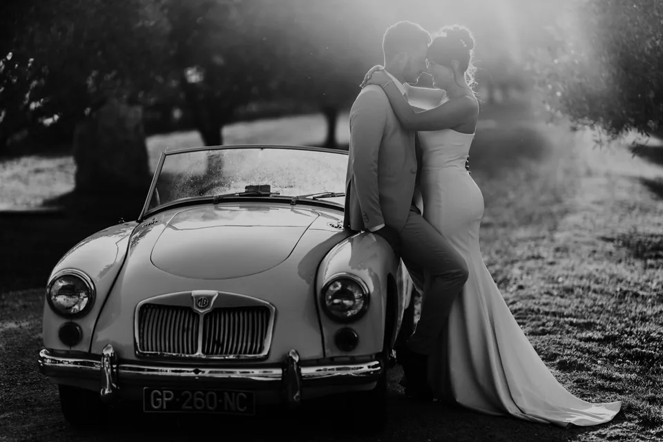 Newlyweds embracing beside a vintage car, in black and white